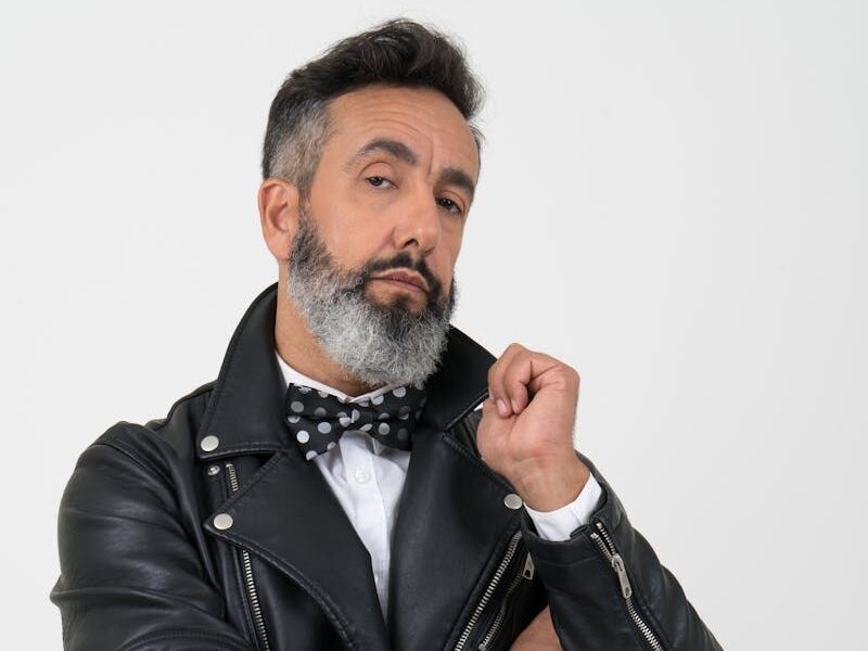 Stylish man posing confidently in a black leather jacket with a bow tie on a white background.