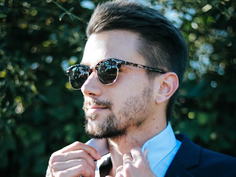 Fashionable man adjusting his suit and sunglasses outdoors on a sunny day.
