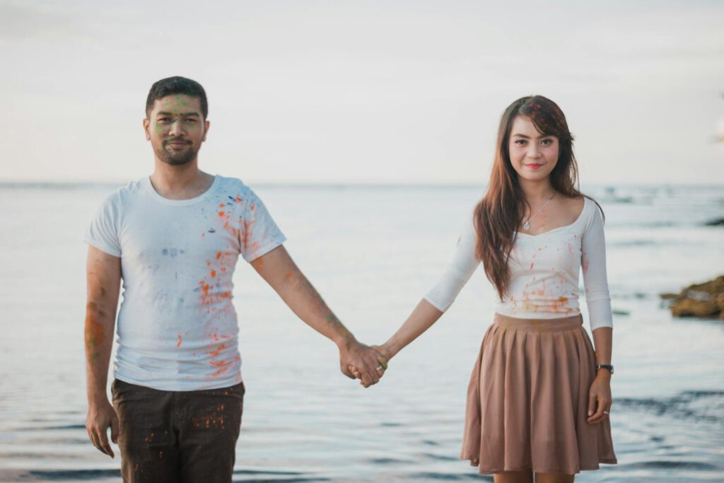 A cheerful couple holding hands by the sea, enjoying a playful day with paint splashes.