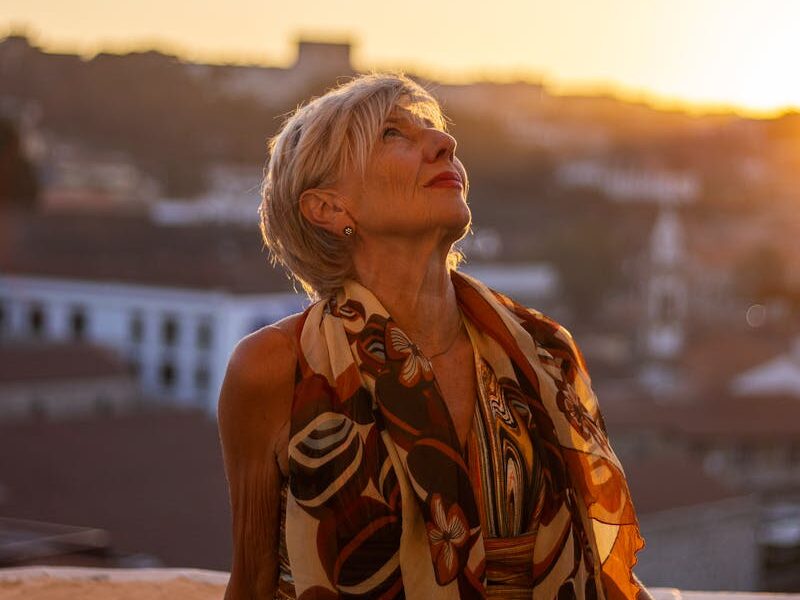 A senior woman in stylish attire looks up during a serene urban sunset, embracing tranquility.
