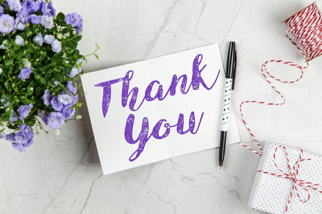 A stylish thank you card paired with purple flowers, a gift box, and pen on a marble surface.