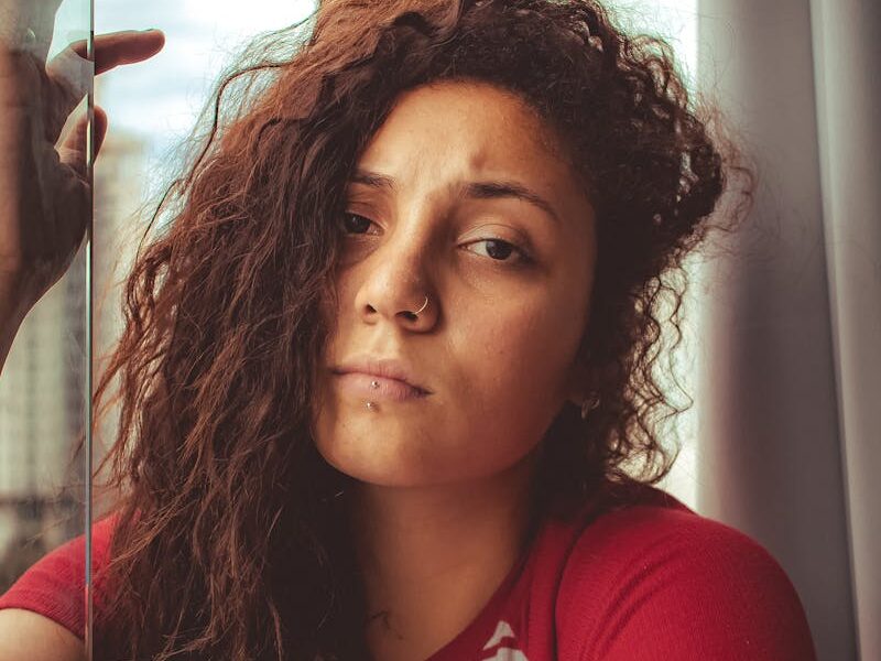 A woman with curly hair looking thoughtfully outside a window in a red striped shirt.