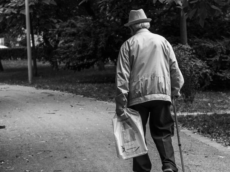 Elderly man in a hat walking on a park path with a cane, captured in monochrome.