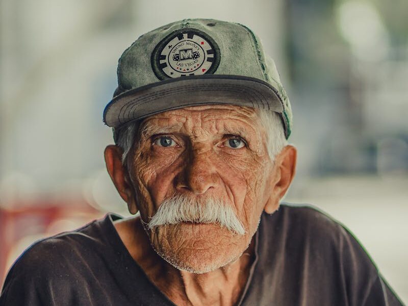 Portrait of an elderly man with a mustache, wearing a cap and serious expression, outdoors.