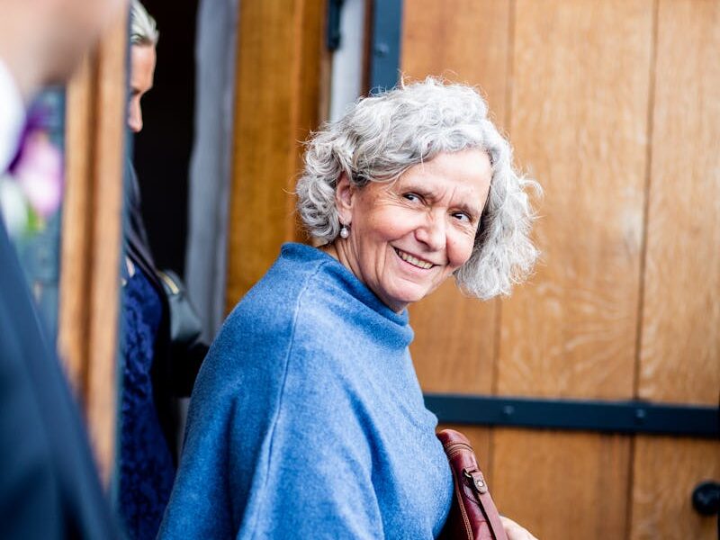 Smiling elderly woman in blue shawl standing at wooden doorway.