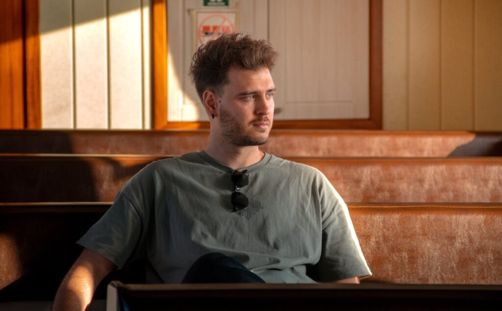 Young man sitting in a sunlit interior, looking contemplative on a wooden bench.