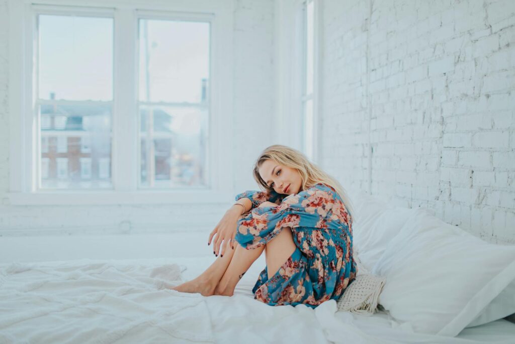 A woman in a floral dress sits on a bed in a modern, brightly lit bedroom, exuding relaxation.