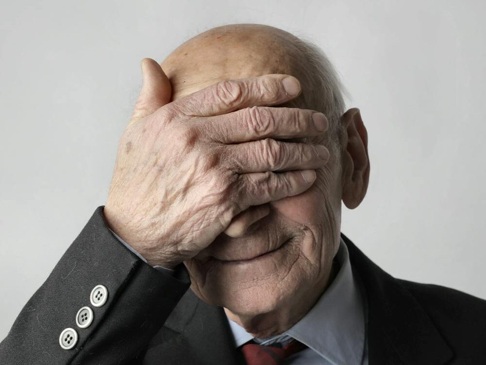 Portrait of a senior man in a suit covering eyes with hand, smiling gently.