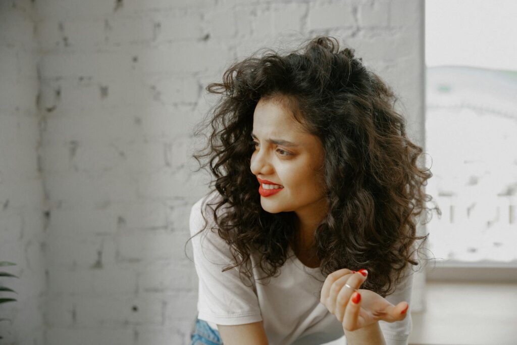 Portrait of a young woman with curly hair showing confusion indoors against a white brick wall.
