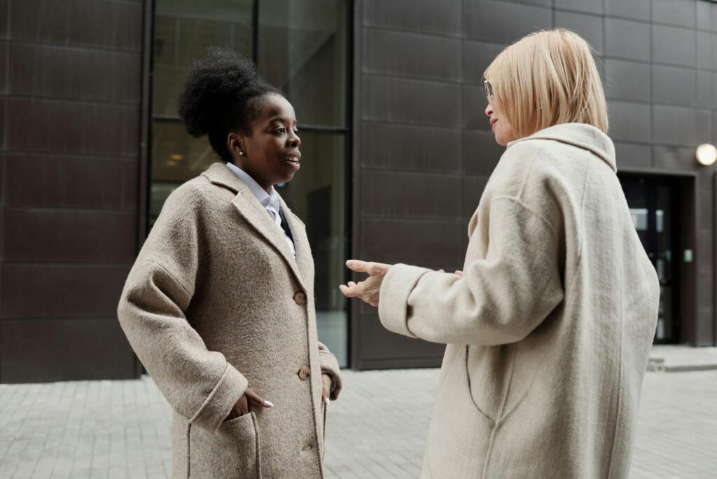 Two businesswomen in coats having a conversation downtown, showcasing professional collaboration.