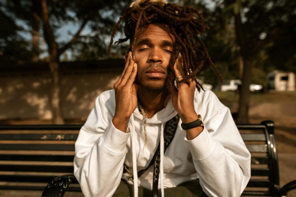 A man with dreadlocks sits on a park bench, contemplating with eyes closed.