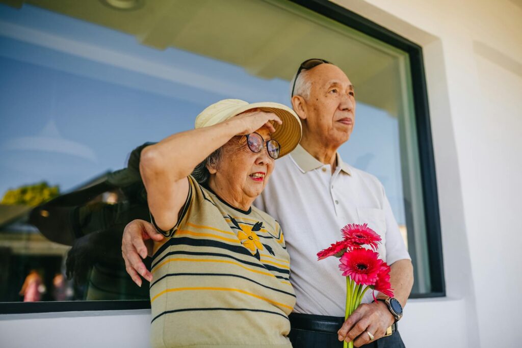 Senior couple standing outside, holding flowers, enjoying a sunny day together.