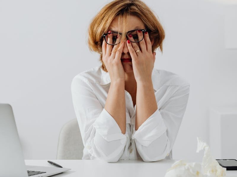 A stressed woman in eyeglasses and white shirt sits at her desk, overwhelmed and exhausted.