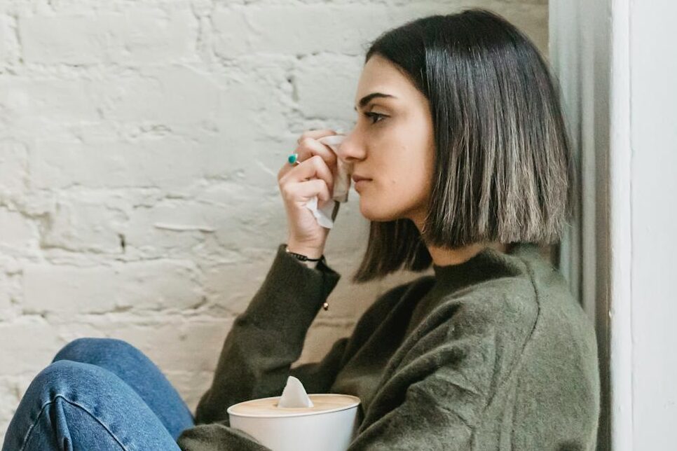 Side view of depressed young ethnic female millennial in warm sweater sitting leaning on wall and wiping tears with tissue at home