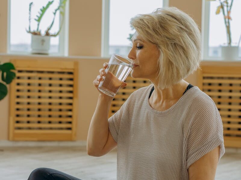 Senior woman in black leggings drinking water while seated on a yoga mat indoors.