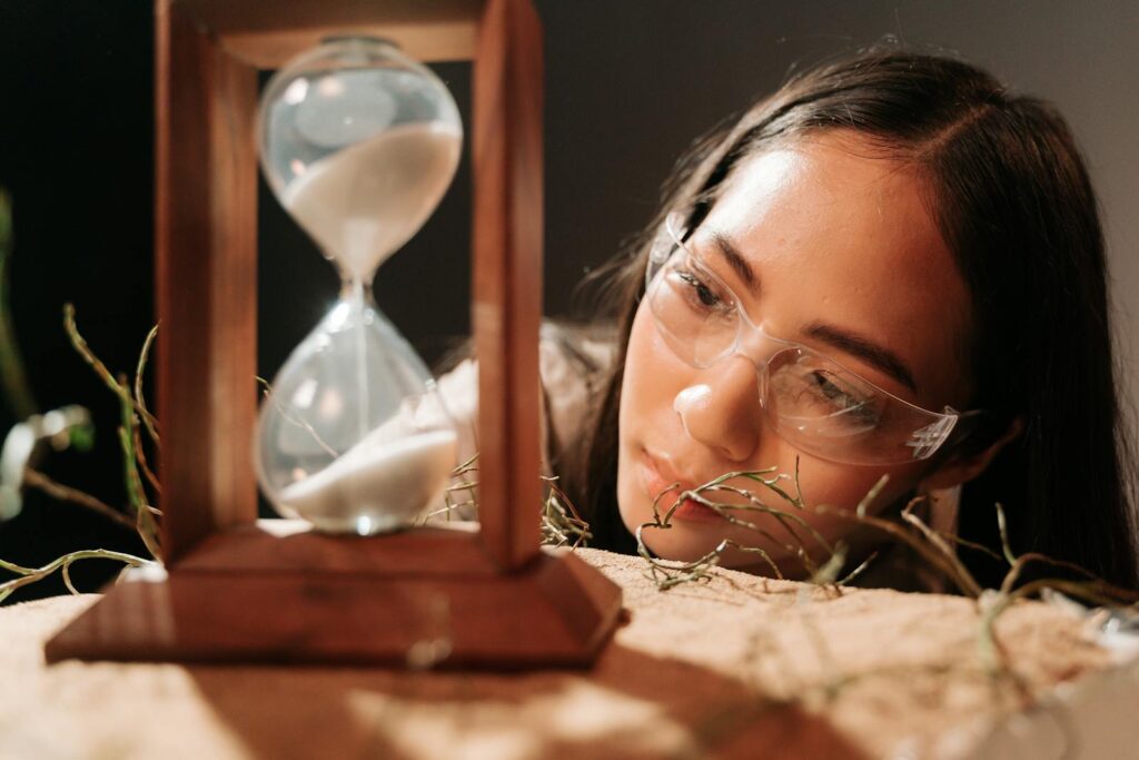 A woman wearing glasses observes an hourglass, symbolizing time and reflection.