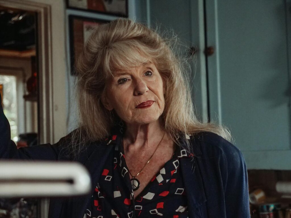 Elderly woman with long blonde hair indoors, looking thoughtful.