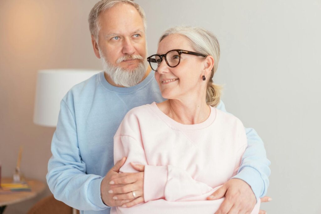 Senior couple in pastel sweaters embracing indoors, showing love and warmth.