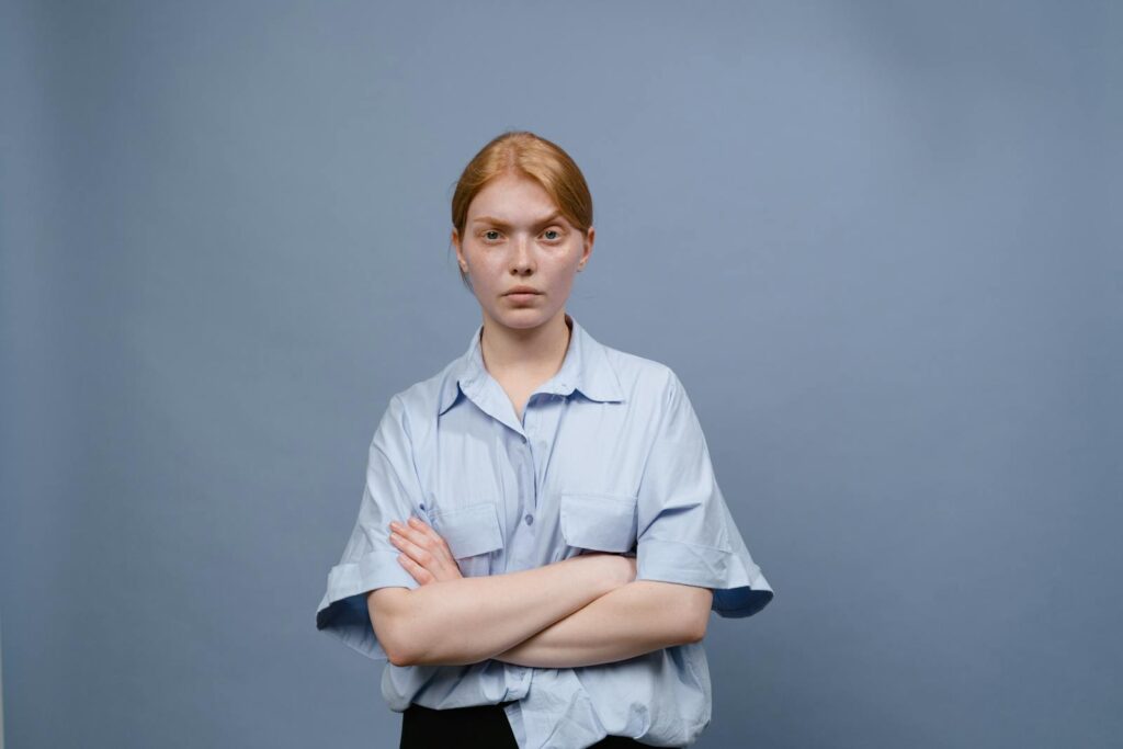 A young woman standing with crossed arms against a plain blue background, expressing annoyance.