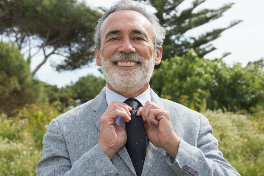 Senior man in suit smiling while adjusting his necktie outdoors against a lush green background.