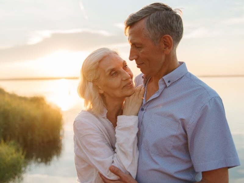 Elderly couple shares a tender moment by a lake during sunset, capturing love and serenity.