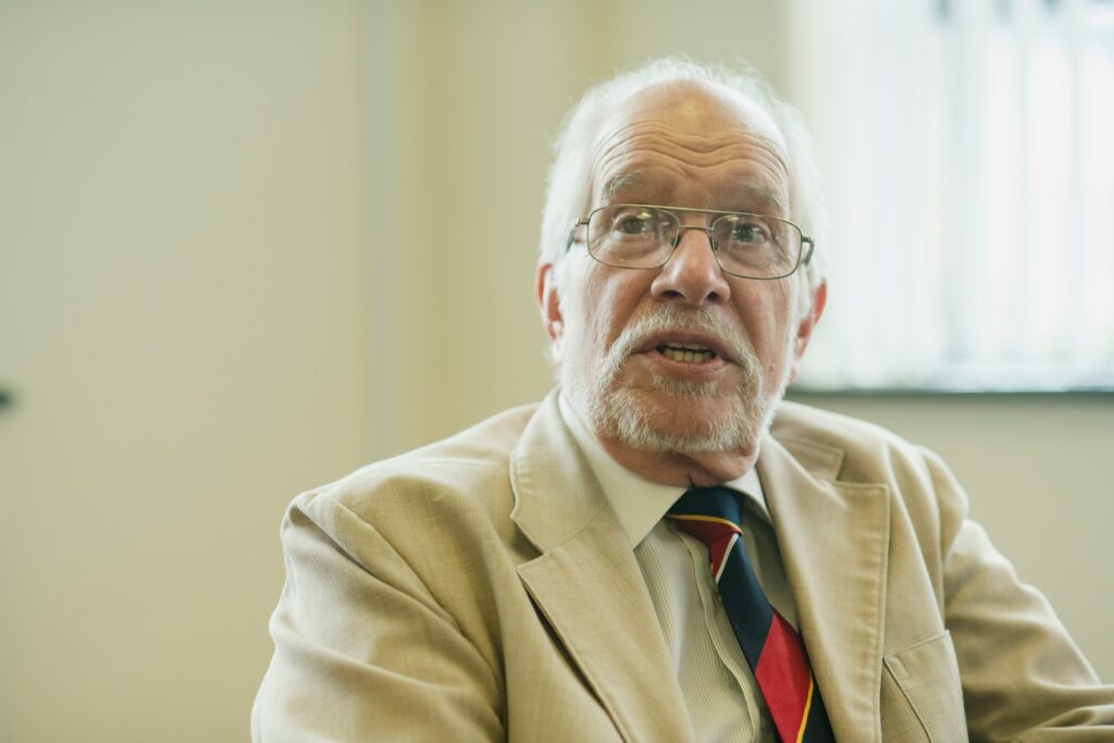 An elderly man in a suit jacket looks at the camera.