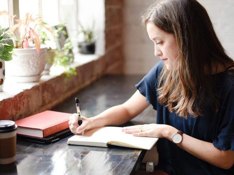 woman sitting in front of black table writing on white book near window