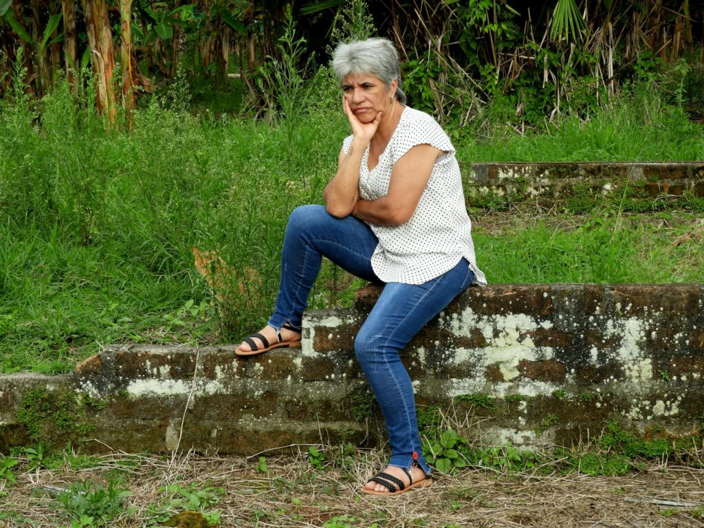 A mature woman in casual attire sits pensively on an old brick wall in a lush outdoor setting.
