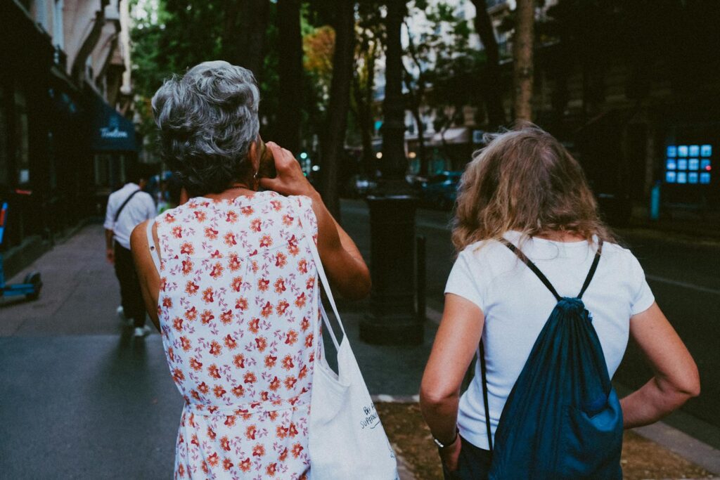 Two women walking on a tree-lined street in an urban setting, captured from behind, illustrating a casual city stroll.