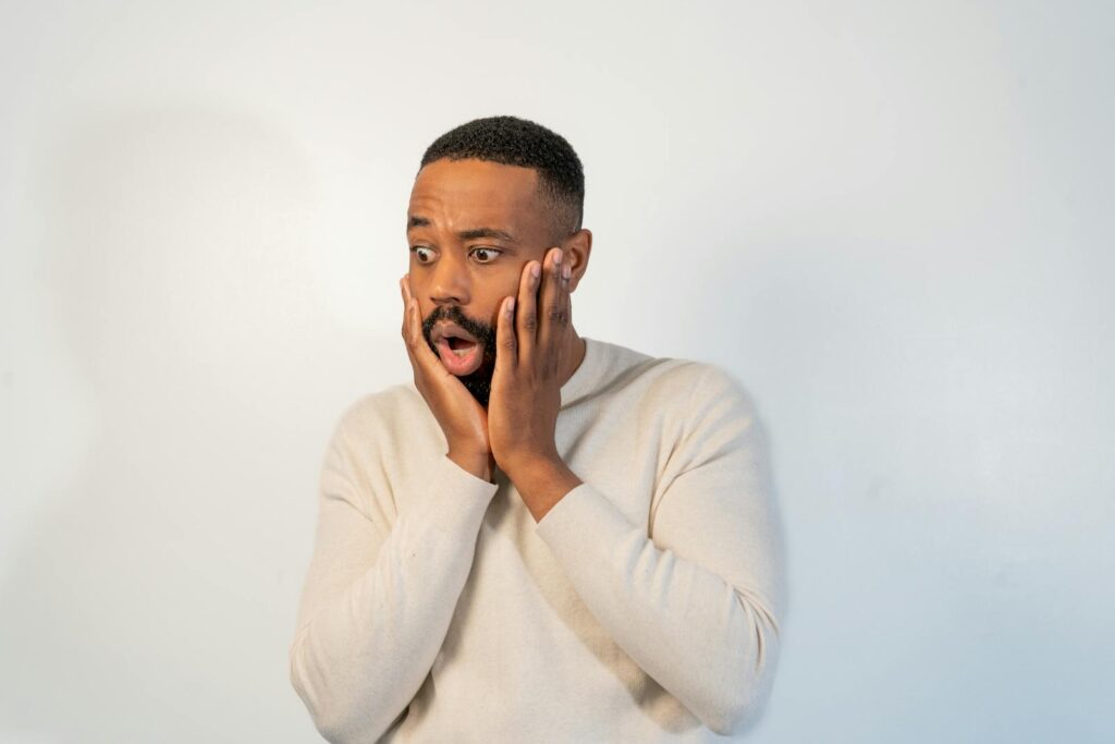 A man expresses shock with hands on face, set against a plain white studio backdrop.