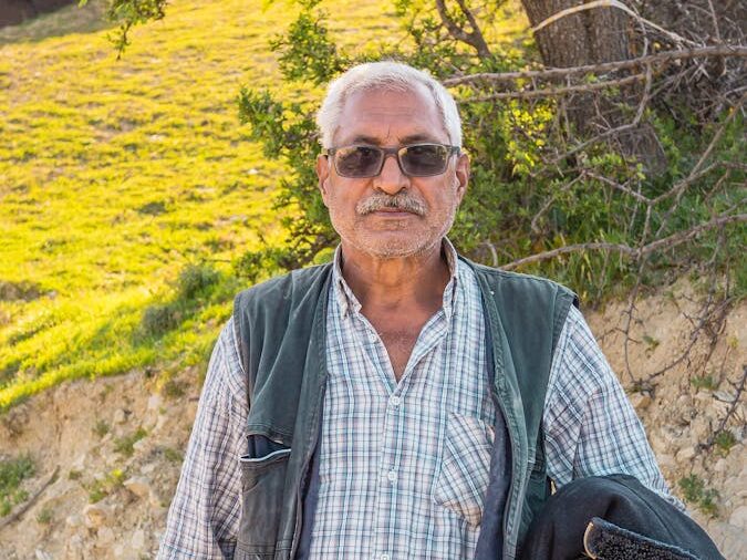 Portrait of an elderly man standing outdoors on a sunny day, in a rural setting.