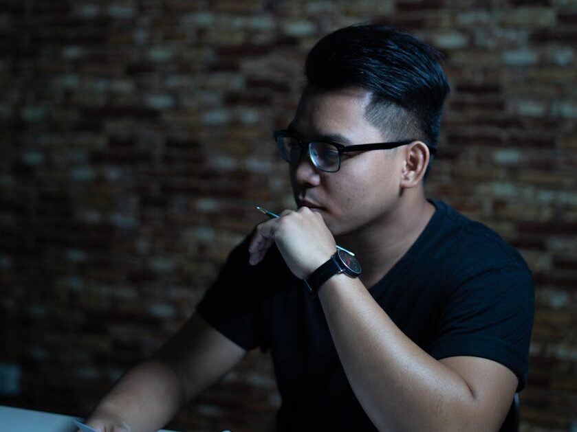 Man wearing glasses thinking with a pen at a table indoors