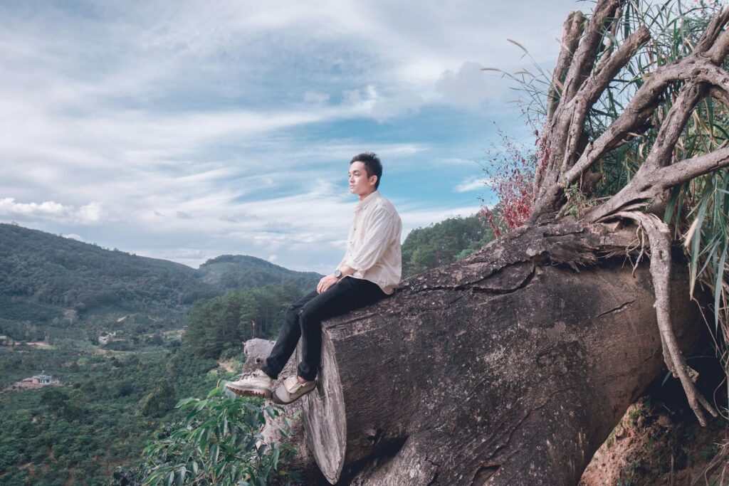 A man sits on a fallen tree overlooking a mountainous landscape, reflecting in nature.