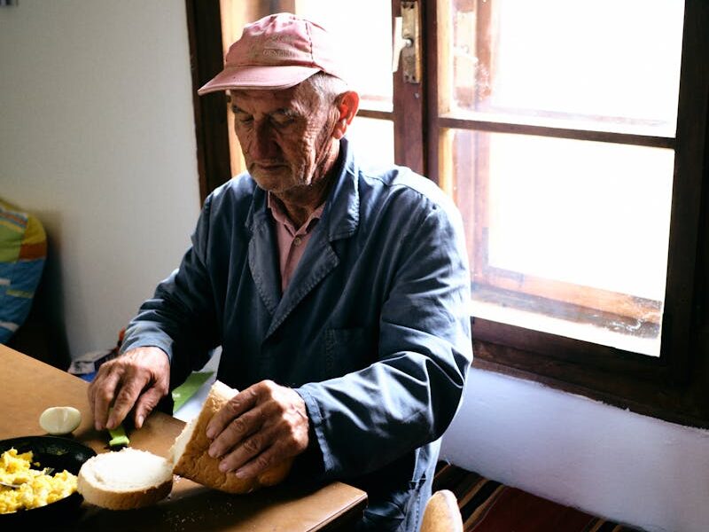 Elderly man in cap sitting at table for breakfast by the window. Cozy natural light setting.