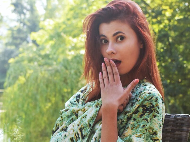 A young woman with red hair displays surprise while seated outdoors amidst greenery.