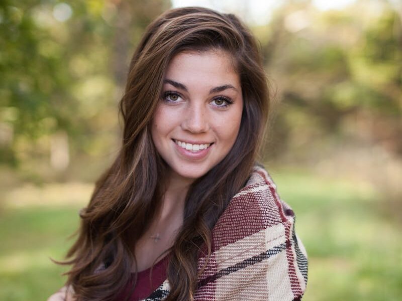 A woman smiling outdoors with a cozy plaid shawl, surrounded by nature in a sunny park.