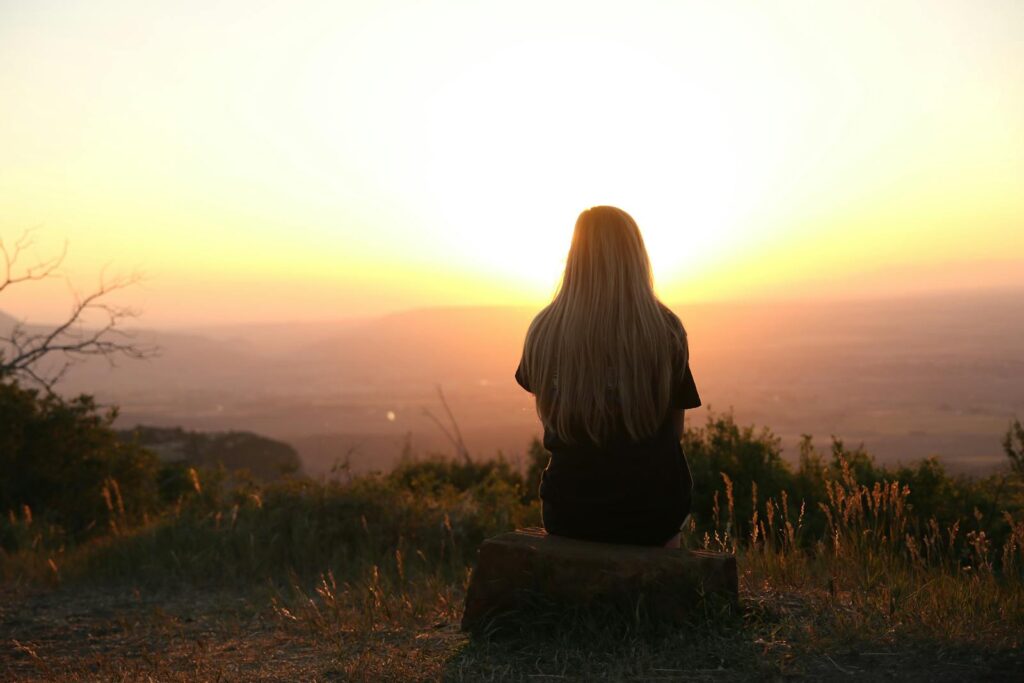 Woman sitting in nature, watching sunset over fields. Peaceful and serene outdoor scene.