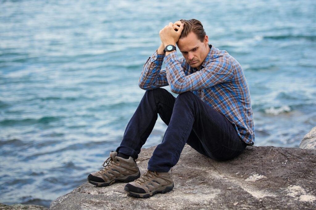 Man in casual wear sitting on a rock by the ocean, looking thoughtful.
