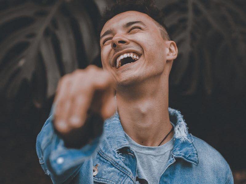 A joyful man in a denim jacket laughs and points, surrounded by lush greenery.