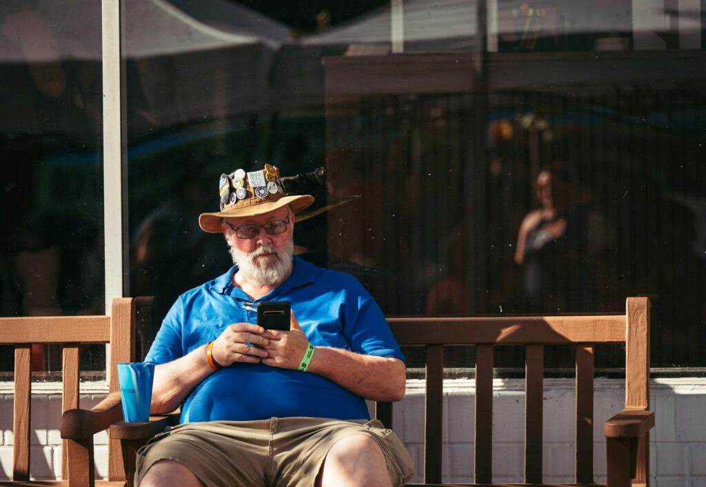 Older man with a decorated hat using smartphone while sitting on a bench outdoors.