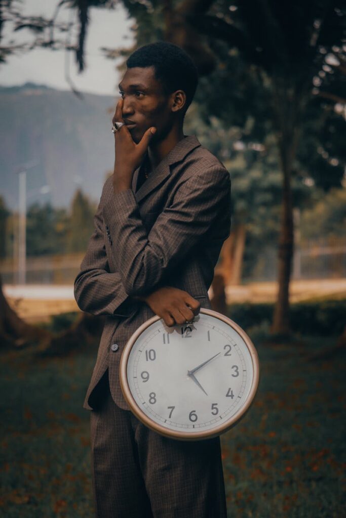 Man in a brown suit outdoors holding a large clock, deep in thought.