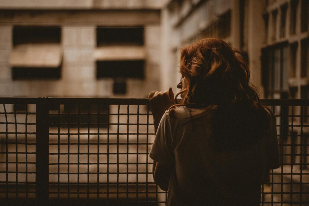 A woman stands alone looking through a railing with a sepia-toned, contemplative mood.