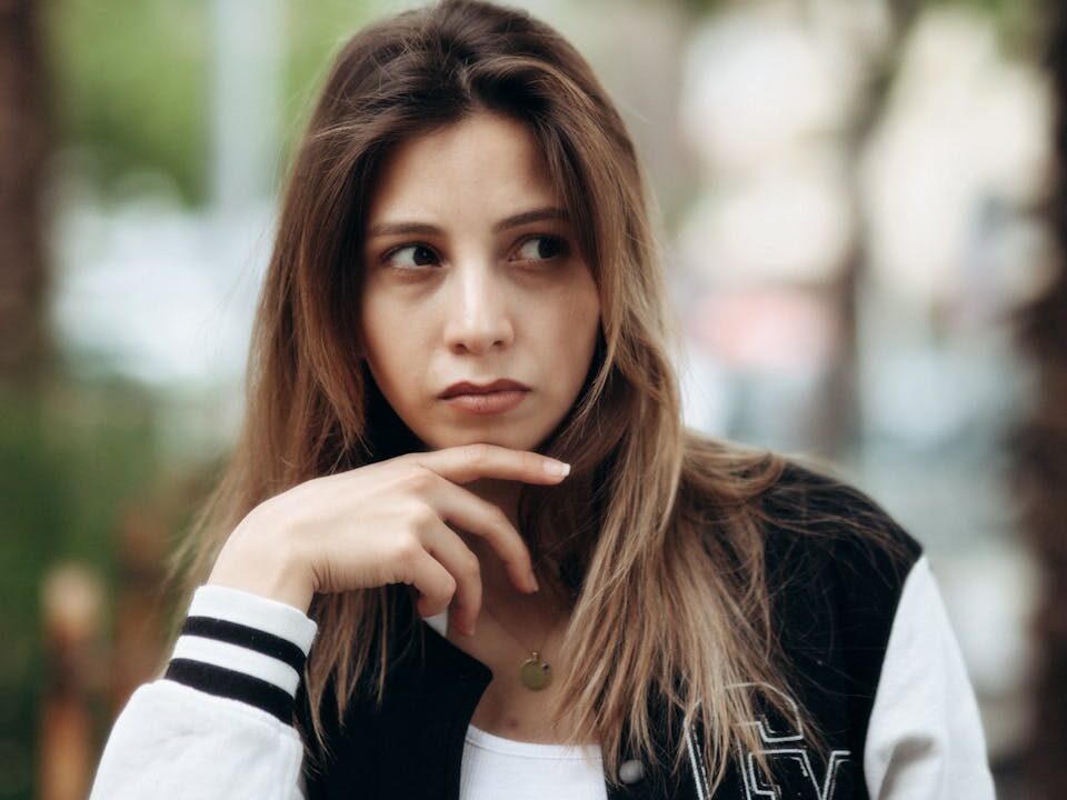 Pensive young adult woman outdoors in a casual jacket, urban backdrop.