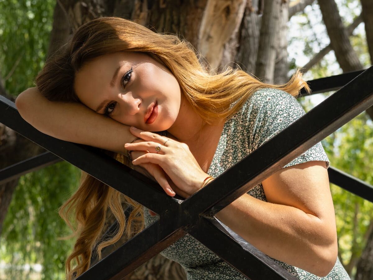 A serene portrait of a woman with long hair leaning on a metal structure in a park. Sun-dappled and peaceful.