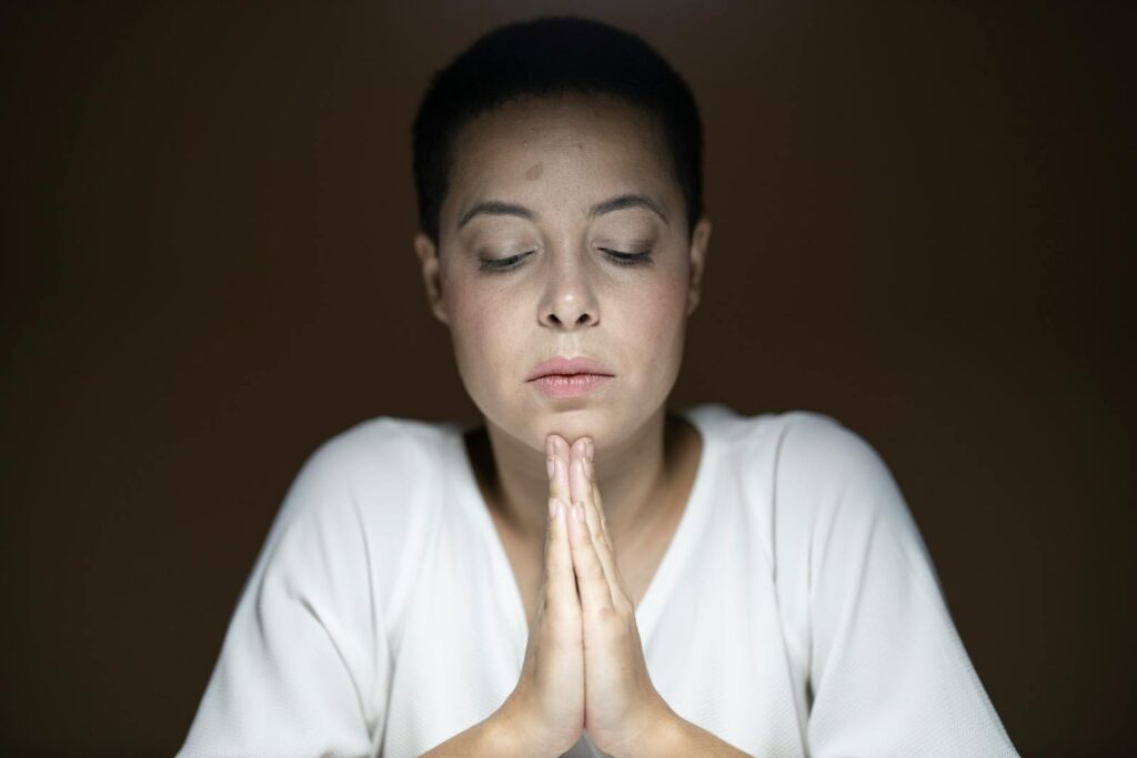 A peaceful woman with closed eyes and clasped hands in deep prayer inside a quiet room.