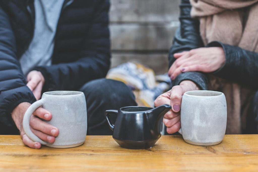 A couple holding mugs of hot beverage with a teapot on a wooden table outside.