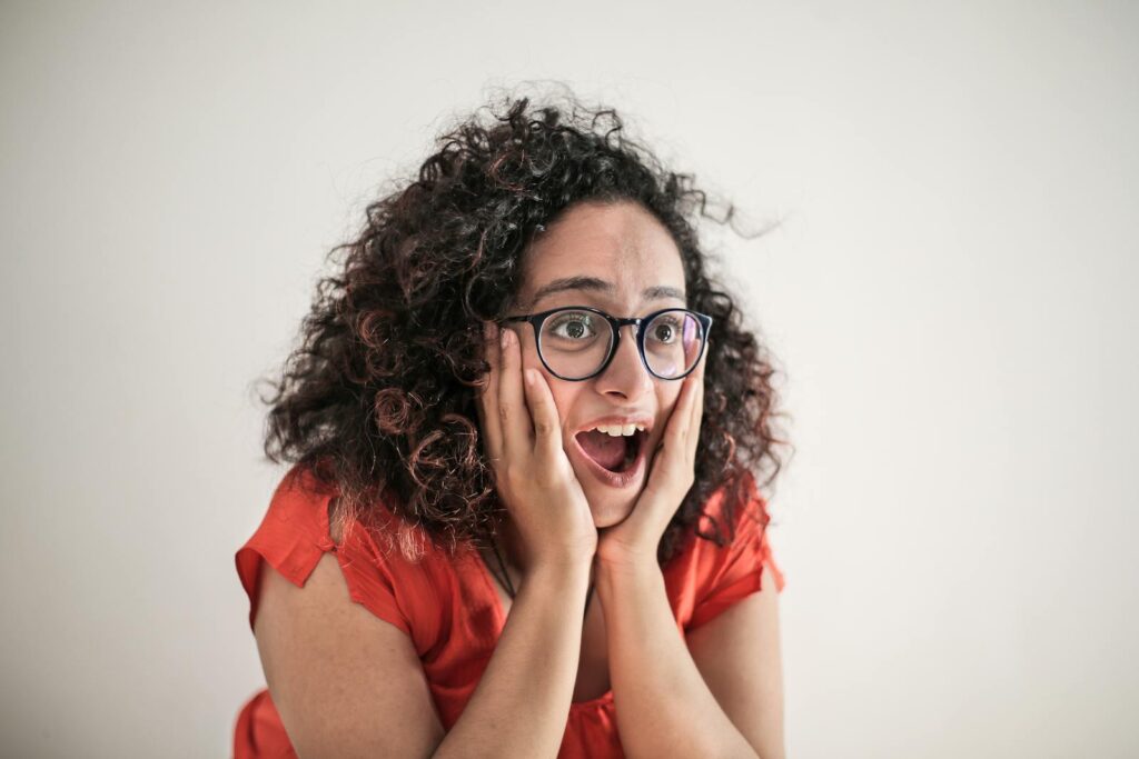 A woman expresses surprise, her curly hair framing her eyeglass-wearing face, hands on cheeks.