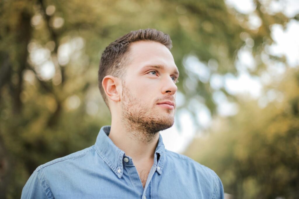 Portrait of a handsome young man with a beard looking thoughtfully in a sunny park setting.