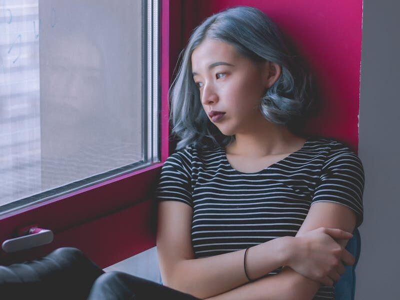 A woman sits by a window, deep in thought, conveying solitude.