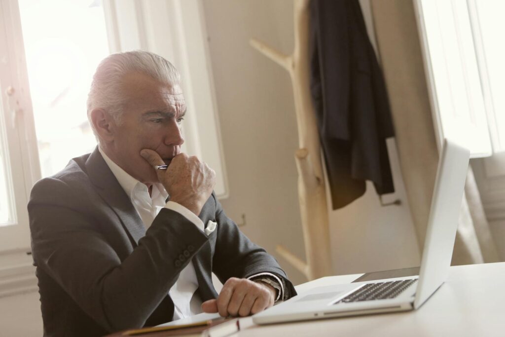 Elderly businessman in a suit thoughtfully working on a laptop at his desk.